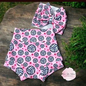 May include: Pink baby shorts and a matching headband with a large bow. Both items are patterned with volleyballs and red stars. The shorts have a high waist design. The headband bow is large and prominent.