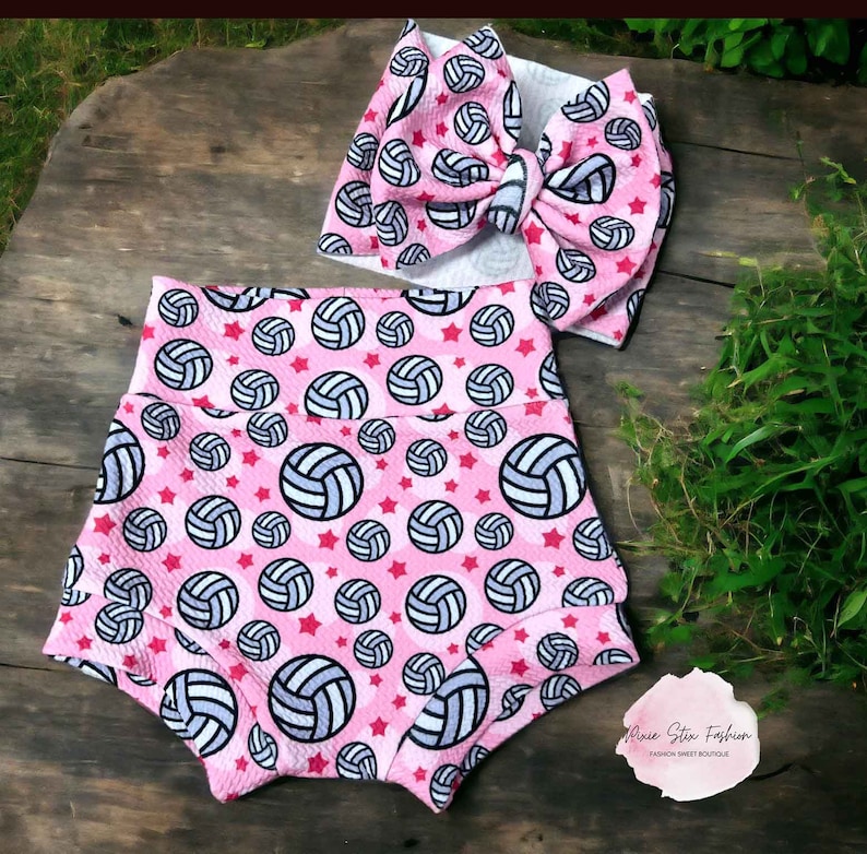 May include: Pink baby shorts and a matching headband with a large bow. Both items are patterned with volleyballs and red stars. The shorts have a high waist design. The headband bow is large and prominent.