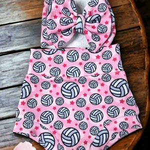 May include: A pink baby outfit set featuring a headband and high-waisted shorts. The set is patterned with volleyballs and red stars. The headband has a large bow. The shorts have a high waistband. The set is on a wooden surface.