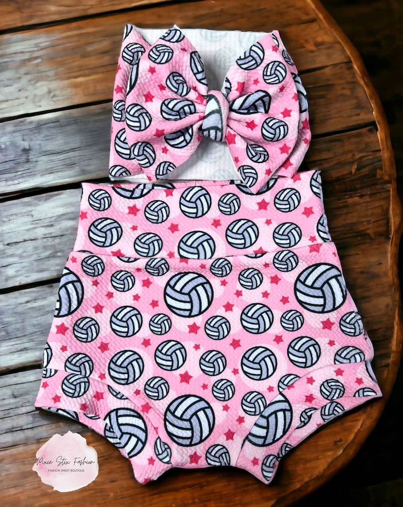 May include: A pink baby outfit set featuring a headband and high-waisted shorts. The set is patterned with volleyballs and red stars. The headband has a large bow. The shorts have a high waistband. The set is on a wooden surface.