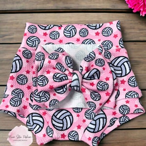 May include: Pink baby shorts and matching bow featuring a volleyball pattern with red stars. The shorts have a high waist design. The bow is large and centered. The volleyballs are white and gray.