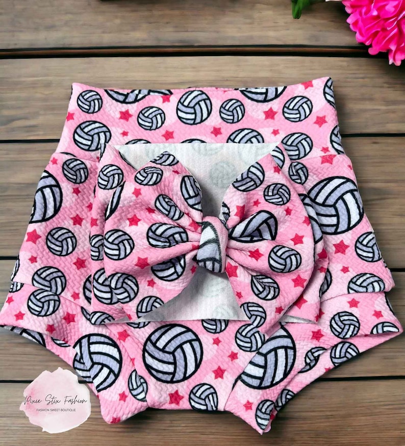 May include: Pink baby shorts and matching bow featuring a volleyball pattern with red stars. The shorts have a high waist design. The bow is large and centered. The volleyballs are white and gray.