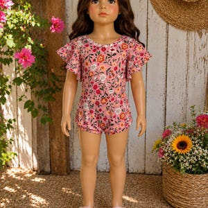 Könnte beinhalten: Ein rosa Blumen-Strampler mit Rüschenärmeln und Shorts, getragen von einer Puppe. Der Strampler hat einen V-Ausschnitt und ein Muster aus roten, orangefarbenen und weißen Blumen. Die Puppe trägt rosa Sandalen mit Blumenakzenten.