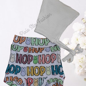 May include: A white baby romper with a colorful 'Hop' pattern and a matching white knit hat. The romper features a repeating pattern of the word 'Hop' and a cartoon bunny with a pastel yellow, green, and pink color scheme.