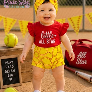 May include: A baby in a red top with "little ALL STAR" text, yellow shorts with baseball stitching, and a matching yellow bow headband. The baby wears white trainers. A sign reads "PLAY HARD DREAM BIG".