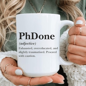 Könnte beinhalten: Weißer Keramikbecher mit dem Text "PhDone" in Schwarz, zusammen mit einer Definition. Der Becher hat einen Henkel und wird von einer Person gehalten. Der Text enthält die Worte "Erschöpft, überqualifiziert und leicht traumatisiert. Vorsicht."