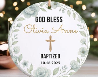 May include: A round, white ceramic ornament with the text "GOD BLESS Olivia Anne" in gold script, a gold cross, and the word "BAPTIZED" below. The date "10.16.2025" is printed below. The ornament is decorated with a floral wreath.