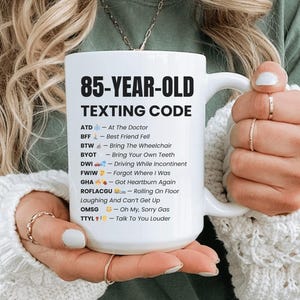 以下が含まれることがあります： 「85-YEAR-OLD TEXTING CODE」の黒い文字がプリントされた白いセラミックマグカップ。マグカップには、「ATD - At The Doctor」や「BFF - Best Friend Fell」など、テキストメッセージの略語とその意味が記載されています。マグカップは人が持っています。