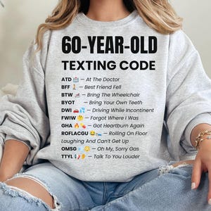 May include: A light grey crewneck sweatshirt featuring the black text 60-YEAR-OLD TEXTING CODE followed by a list of humorous acronyms and emojis, including ATD, BFF, BTW, BYOT, DWI, FWIW, GHA, ROFLACGU, OMSG, and TTYL with their definitions.