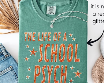 May include: A teal Comfort Colors t-shirt with the text "THE LIFE OF A SCHOOL PSYCH" in orange glitter lettering. The shirt is folded and accessorized with a silver necklace. Denim jeans and a beige scarf are visible in the background.