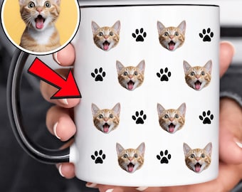 May include: White ceramic mug with a black rim and handle, decorated with repeating images of a tabby cat's face and black paw prints. A circular image of the cat is in the upper left corner. The cat has its mouth open.
