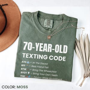 May include: A moss green t-shirt with the text "70-YEAR-OLD TEXTING CODE" in white. The shirt includes a list of texting abbreviations and their meanings. A gold necklace and a brown hat are also visible.