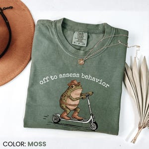 Pode incluir: Camiseta verde musgo com a frase "off to assess behavior" e um sapo de desenho animado com chapéu e botas de cowboy numa scooter. Um colar de ouro e um chapéu castanho também são visíveis.