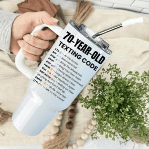 May include: A white and silver insulated tumbler with a handle, featuring the text "70-YEAR-OLD TEXTING CODE" in black. The tumbler includes a straw and a list of texting acronyms. The tumbler is held by a person.