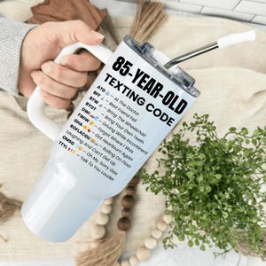 May include: A white and silver insulated tumbler with a white handle and a straw. The tumbler features the text "85-YEAR-OLD TEXTING CODE" in black, along with a list of texting acronyms and their meanings.