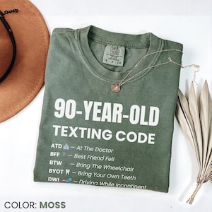 May include: Moss green t-shirt with the text "90-YEAR-OLD TEXTING CODE" in white. The shirt lists texting abbreviations and their meanings. A gold necklace and a brown hat are also visible.