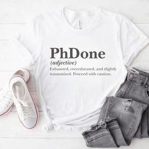 May include: White t-shirt with a black graphic that says "PhDone (adjective) Exhausted, overeducated, and slightly traumatized. Proceed with caution."