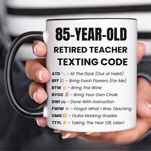 Puede incluir: Taza de cerámica blanca con asa y borde negros. La taza presenta el texto "85-YEAR-OLD RETIRED TEACHER TEXTING CODE" y una lista de abreviaturas de mensajes de texto. Un regalo divertido para un profesor jubilado.