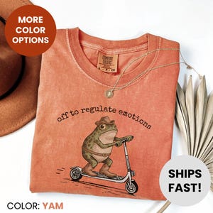 Op de afbeelding: Een yam-kleurig t-shirt met de tekst "off to regulate emotions" en een afbeelding van een kikker met een cowboyhoed op een scooter. Het shirt heeft een ronde hals en korte mouwen. De tekst "MORE COLOR OPTIONS" en "SHIPS FAST!" zijn ook zichtbaar.