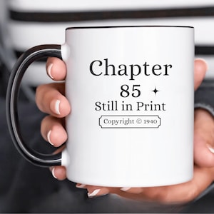 Op de afbeelding: Witte keramische mok met een zwarte handgreep en rand. De mok heeft de tekst "Chapter 85 Still in Print" in een vetgedrukte, zwarte letter. Daaronder staat "Copyright © 1940" in een kader. De mok wordt door een persoon vastgehouden.