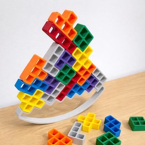 Pode incluir: Um brinquedo de baloiço de madeira multicolorido feito de blocos interligados em várias cores, incluindo laranja, vermelho, verde, amarelo, azul e roxo. O brinquedo foi projetado para balançar sobre uma base cinza. Vários blocos soltos estão espalhados.