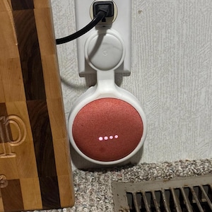 May include: A white and coral smart speaker plugged into a wall socket. The speaker has a fabric-covered front and four illuminated dots. A wooden chopping board is visible on the left.