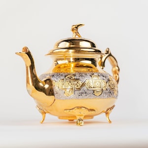 Puede incluir: Una tetera dorada con intrincados diseños de filigrana plateada y dorada. La tetera tiene un pico curvo, un asa decorativa y una tapa con un adorno en forma de pájaro. Se apoya sobre tres pequeños pies.