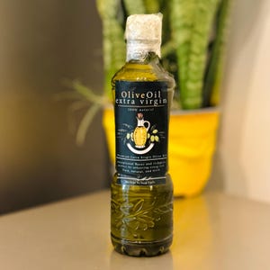 May include: A glass bottle of olive oil with a black label that reads "Olive Oil extra virgin 100% natural". The label also features a drawing of a jug of olive oil and olives.