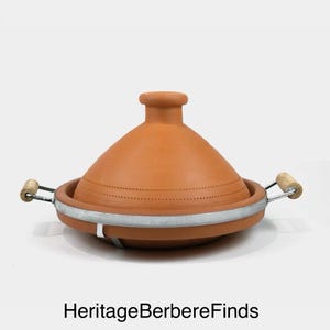 May include: A terracotta tagine pot with a conical lid and a circular base. The pot has a metal band around the base with wooden handles. The text "HeritageBerbereFinds" is at the bottom.