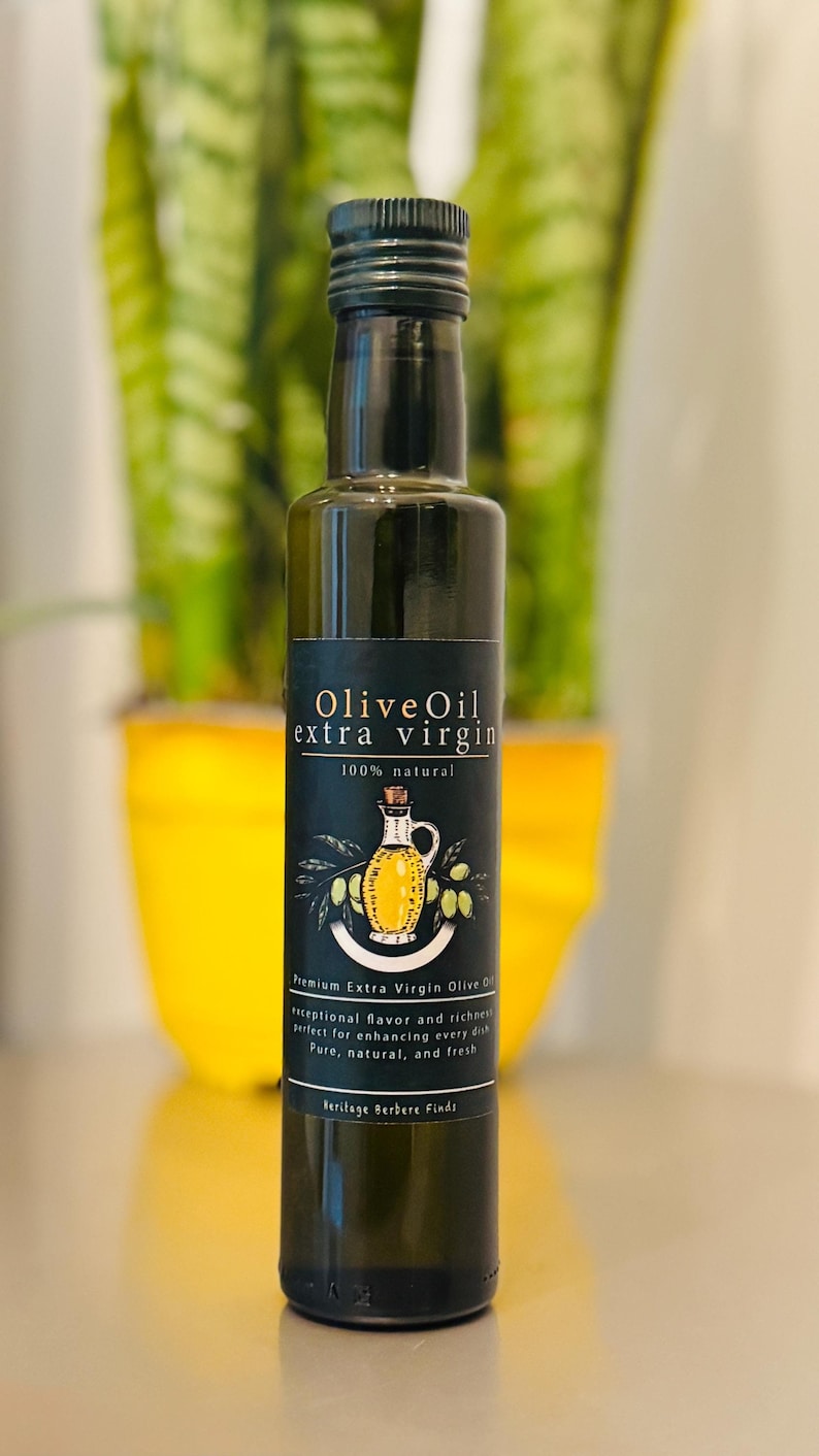 May include: A glass bottle of olive oil with a black label that reads "Olive Oil extra virgin 100% natural". The label also features a drawing of a pitcher of olive oil and olive branches. The bottle is sitting on a white surface.