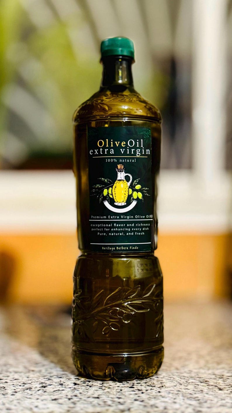 May include: A glass bottle of 100% natural extra virgin olive oil with a green label that reads "Olive Oil extra virgin 100% natural". The label also features a drawing of a traditional olive oil jug and olives.