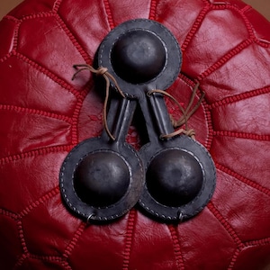May include: Three black castanets with leather ties rest on a red leather ottoman. The castanets have a circular shape with a raised center and a decorative stitched edge. The ottoman has a geometric pattern.