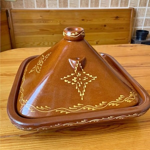 Puede incluir: Una olla de tagine de cerámica marrón con tapa cónica y base cuadrada. La tapa está decorada con motivos florales amarillos y un diseño de estrella. La olla está sobre una mesa de madera.