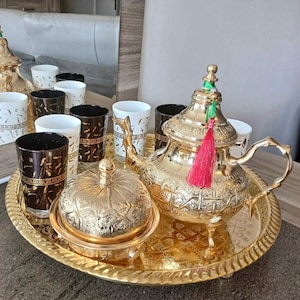 May include: A gold tea set comprising a teapot, sugar bowl, and glasses on a matching tray. The teapot features a red tassel and green details. The glasses alternate between white and brown with decorative patterns.