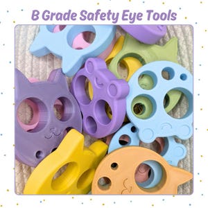 May include: A collection of colorful, plastic safety eye tools in various shapes, including cats, fish, and circles. The tools are in shades of purple, yellow, blue, and green. The text "B Grade Safety Eye Tools" is at the top of the image.