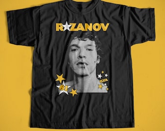 May include: Black t-shirt featuring a grayscale portrait of a person smoking a cigarette. The shirt has the name "RAZANOV" in yellow block letters above the portrait, with yellow star graphics and the text "ILYA" and "81" in yellow.