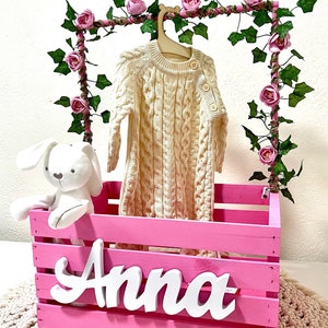 May include: Pink wooden crate with the name "Anna" on the front. The crate is decorated with a garland of pink flowers and green leaves. A white knitted baby outfit hangs on a wooden hanger inside the crate. A white stuffed bunny sits in the crate.