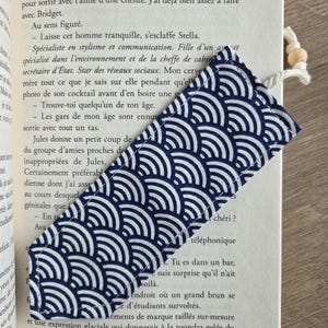 May include: A navy blue and white patterned fabric bookmark is inserted into a book. The bookmark features a repeating wave design. A white cord with a wooden bead is attached to the top of the bookmark.