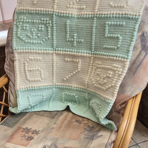 May include: A crocheted baby blanket with a light green and cream color scheme. The blanket has a textured design with raised dots and the word "LOVE" is spelled out in the center.