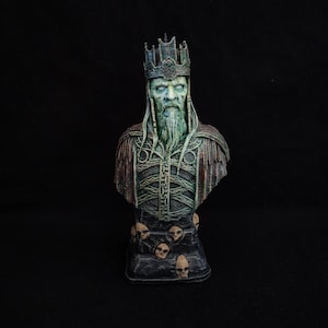Può includere: Busto dettagliato di un re con corona, lunga barba e armatura ornata. La scultura ha un aspetto invecchiato con toni verdi e marroni. La base presenta dettagli di teschi, aggiungendo un tema gotico o fantasy.