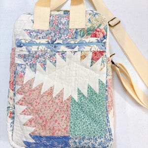 May include: A quilted crossbody bag with a cream-colored adjustable strap and top handle. The bag features a patchwork design with floral patterns in pink, blue, and green, and a light blue zipper. The bag is made of fabric.