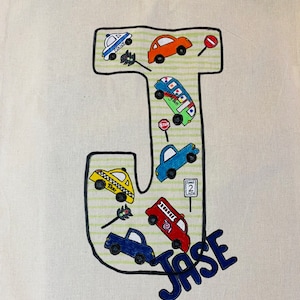 May include: A white canvas tote bag with a large black letter J decorated with colorful cartoon cars and the name "Jase" in blue letters.