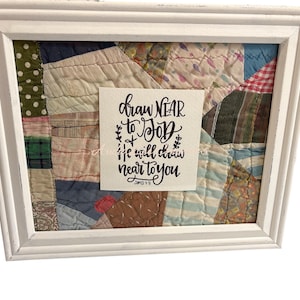 May include: A framed patchwork quilt with a white distressed wooden frame. A small white card in the center displays the handwritten text "draw NEAR to God, he will draw near to you" from James 4:8.