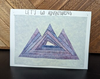 May include: A white card with the text "LET'S GO ADVENTURING" printed at the top. The card features a layered, geometric mountain design in shades of purple and blue on a light blue background.