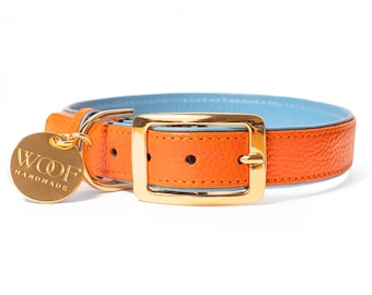 Puede incluir: Un collar de color naranja brillante con textura y forro interior azul claro. Cuenta con una hebilla de metal dorado pulido y una etiqueta redonda de metal dorado que cuelga de una anilla, grabada con el texto WOOF HANDMADE en mayúsculas.