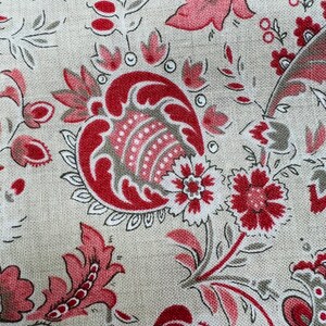 May include: Close-up of a fabric with a repeating floral pattern. The design features red and pink flowers, paisley shapes, and leaves on a beige background. The fabric has a textured appearance, suggesting a woven material.