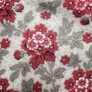 May include: Close-up of a fabric with a floral pattern. The design features clusters of red flowers with white centers and smaller red blossoms. Gray-green leaves and stems are interspersed on a beige background, creating a vintage aesthetic.