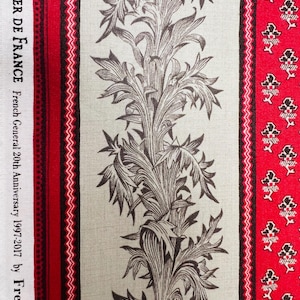 May include: Close-up of a fabric with a repeating floral pattern in brown and white on a light green background. The fabric has red and white striped borders with text that reads "ATELIER DE FRANCE French General 20th Anniversary 1997-2017 by French Ge".
