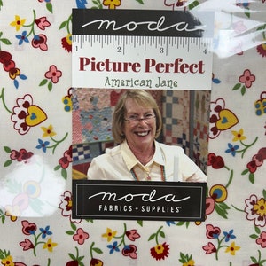 May include: White fabric with a floral pattern in red, pink, yellow, and blue. The fabric has a label that reads "Picture Perfect American Jane" and "moda FABRICS + SUPPLIES". A woman is pictured on the label.