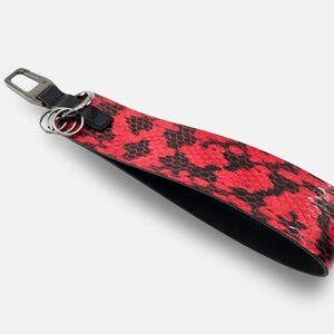 以下が含まれることがあります： 赤と黒のヘビ柄のリストストラップ。金属製の留め金とキーリングが付いています。ストラップは質感のある素材で作られています。金具はシルバーカラーです。手首に巻いて使用するように設計されています。